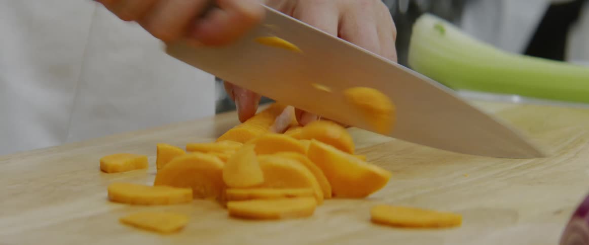 A male quickly cuts a carrot into slices on a cutting board using a large kitchen knife