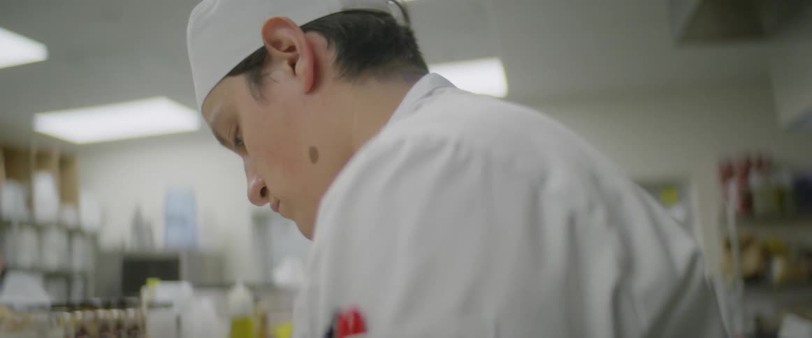 A male chef in a commercial kitchen is looking down while preparing food