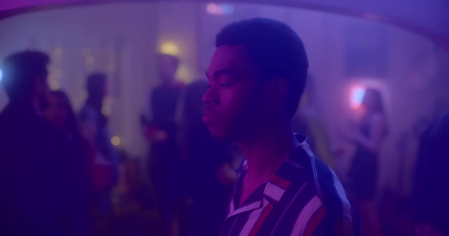 A man walks through a house party where people are drinking and dancing