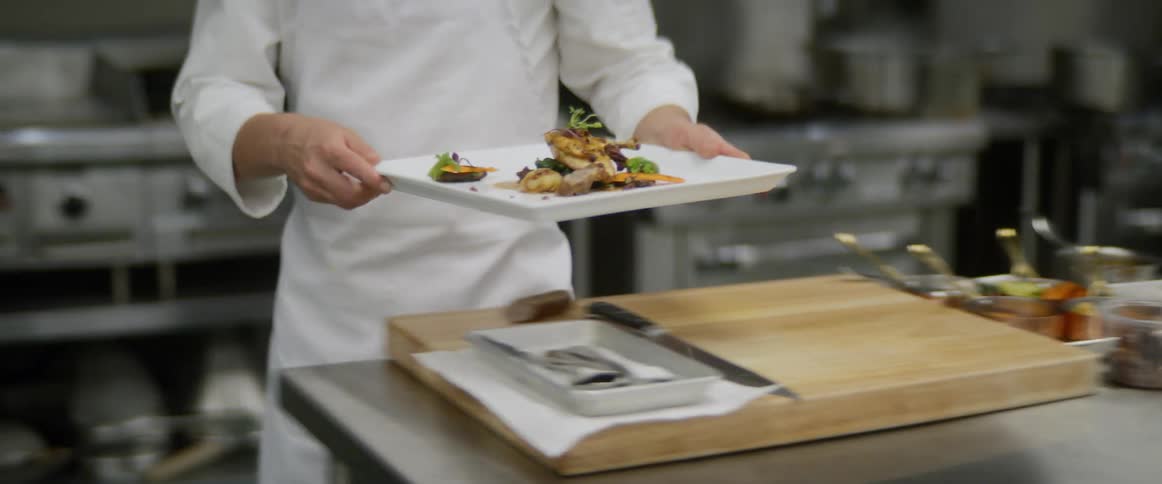 A male chef carries an elegantly prepared plate of food and sets it on a stainless steel countertop