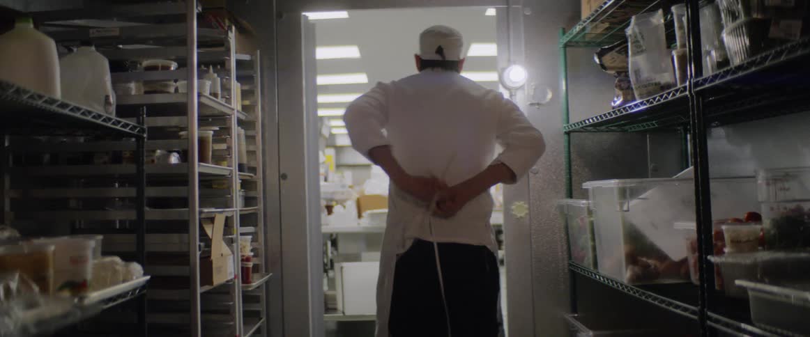 A man in a walk in refrigerator tying his apron and walking out