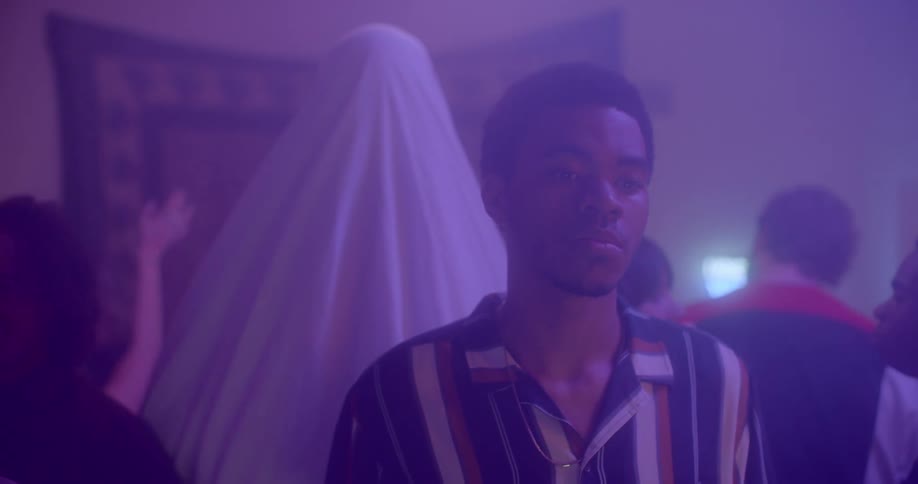 A young black teenager stands alone at a party as a person in a white sheet walks up behind him