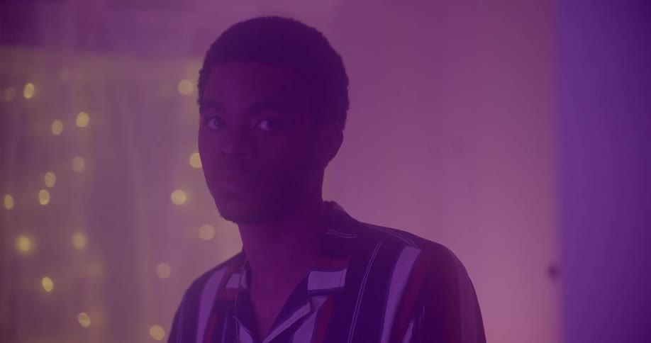 A man in a striped shirt stands and stares in a bedroom with fairy lights
