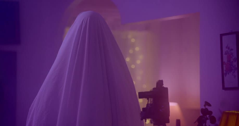 A person with a white sheet completely covering them stands in a room while people dance and party