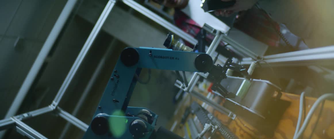 Two men standing and talking in front of a robot arm and a conveyor belt