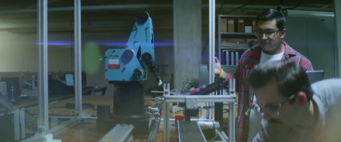 Two men looking at a robotic arm while one is holding a laptop