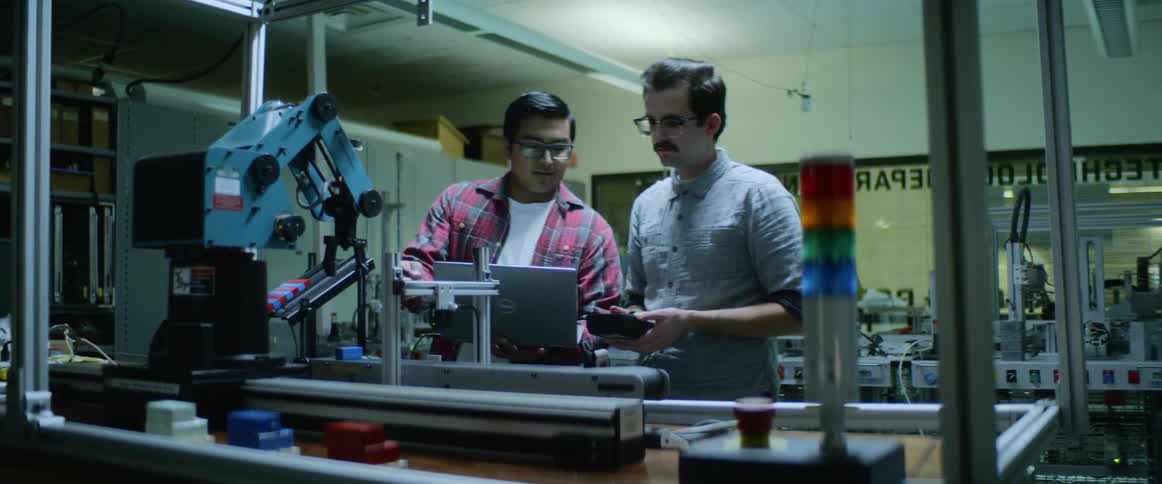 Two men look at a laptop connected to a 3d printer in a workshop