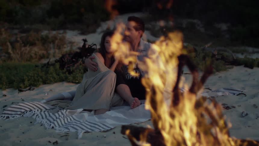Man and woman sitting in sand embracing near a campfire