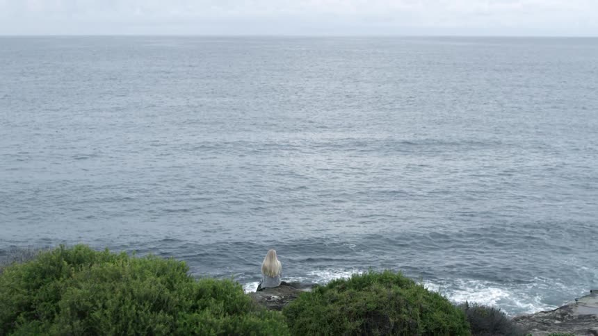 A woman with blond hair sits on a rock next to the ocean