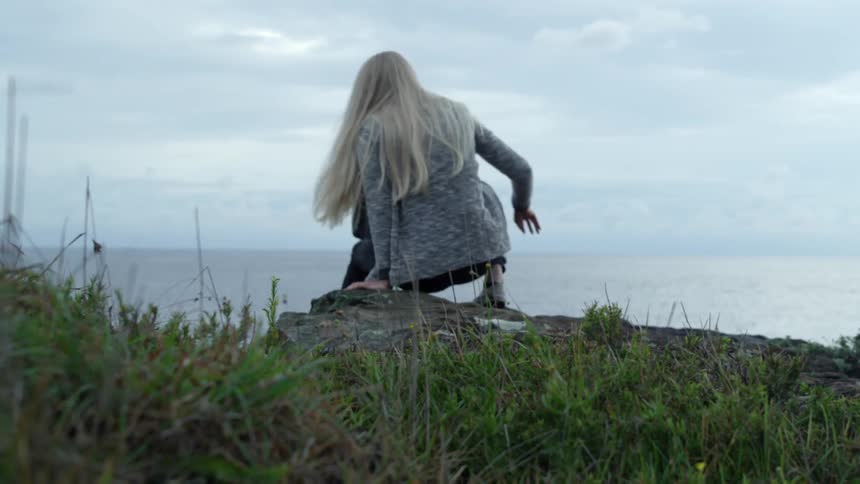 A woman walks to the edge of a cliff and sits down on a cloudy day