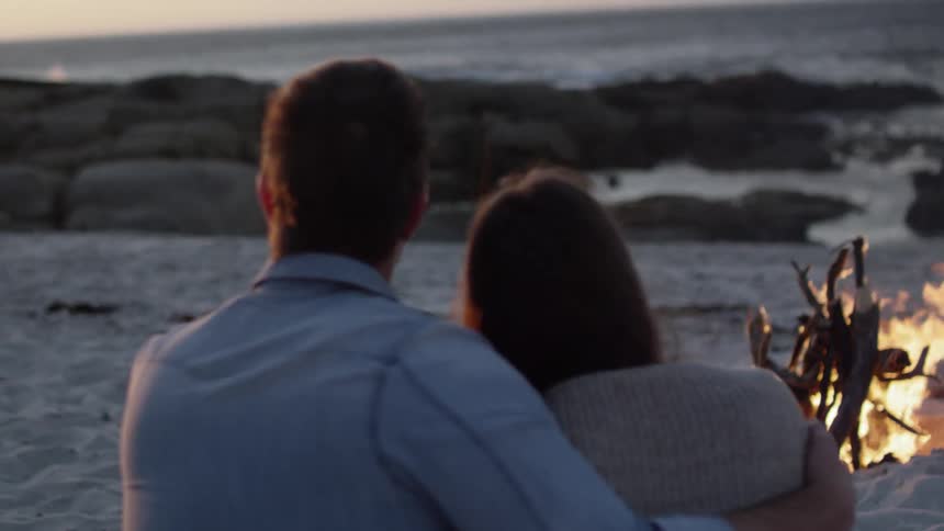 Man and woman sitting in sand embracing near a campfire