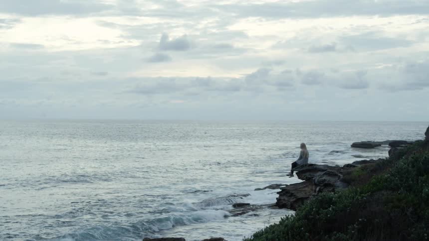 A woman sits on a rock overlooking the ocean on a cloudy day