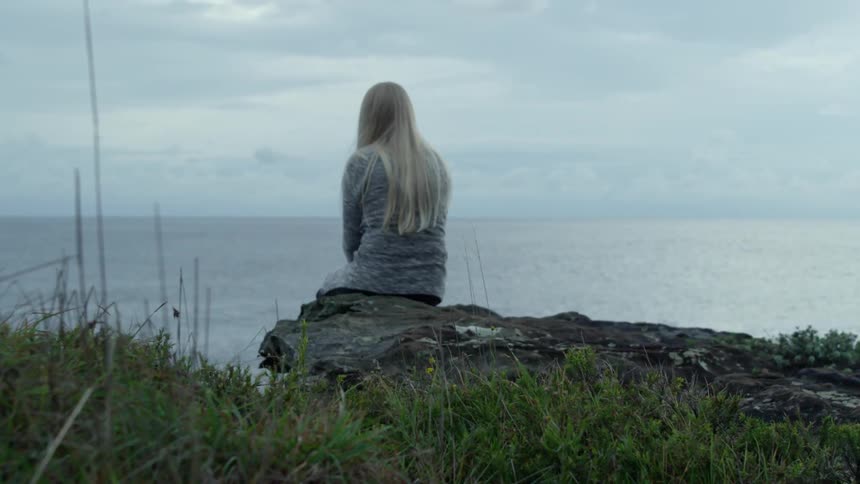 A woman is sitting on the edge of a cliff by the water on a cloudy day