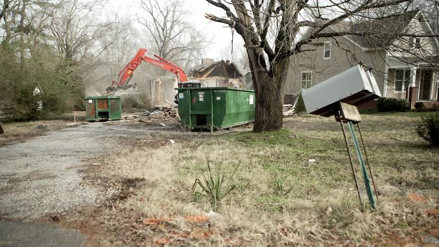A crane at a house loads debris in to a big green dumpster