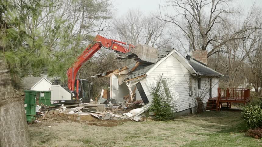 An excavator knocks down a small white house in a neighborhood