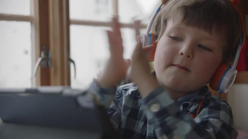 A young boy wearing headphone looks at a computer tablet screen and claps his hands