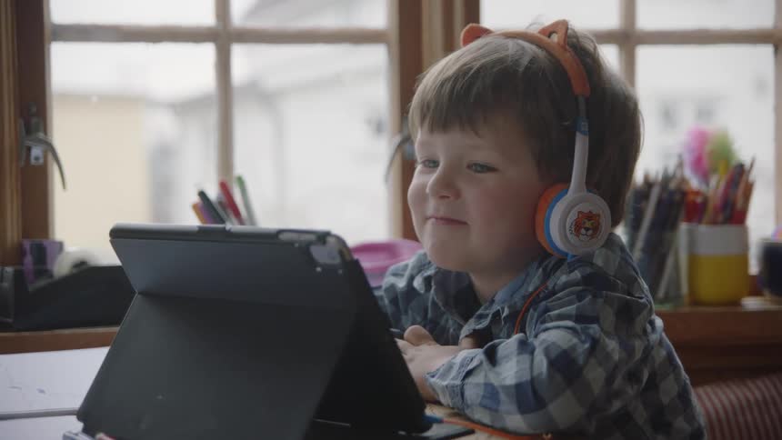 A young boy is sitting at his desk at school watching a table while smiling and wearing tiger headphones