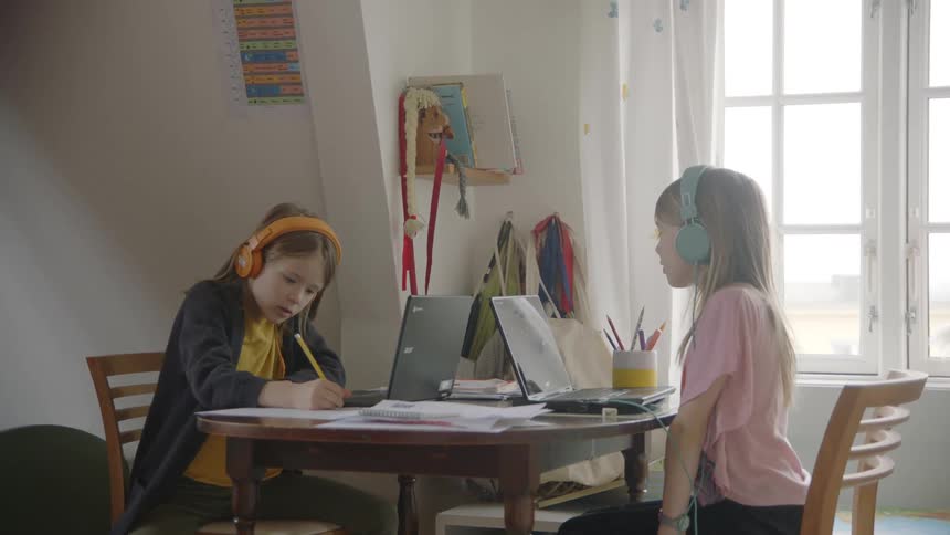 Two girls wearing headphones using laptops on a small table