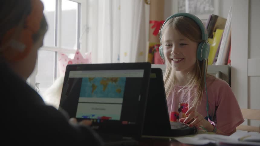 A young girl sits in front of a laptop while smiling and wearing headphones