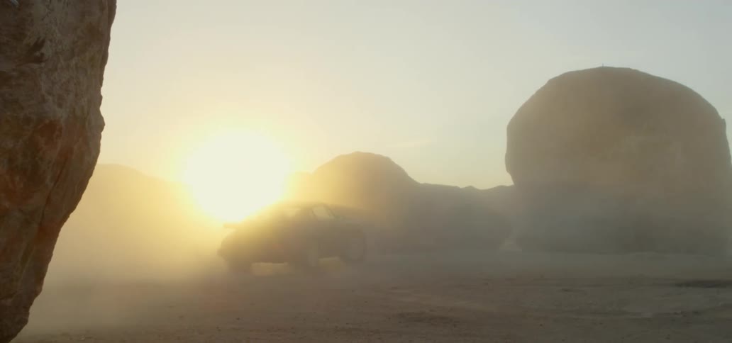 Sun glares through the dust kicked up by car driving among large boulders