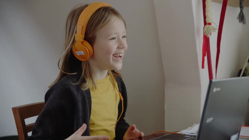 Little girl sitting at the table wears orange headphones and uses the laptop in her room