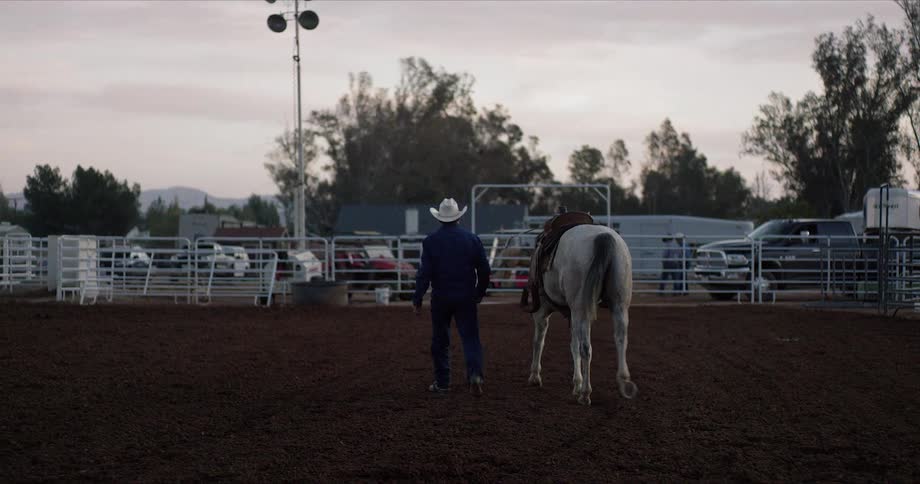 Man an saddled horse walk across padlock toward fence after dusk