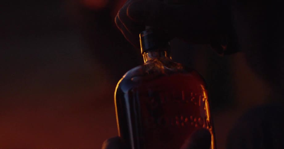 Person opens liquor bottle outdoors near bonfire at night