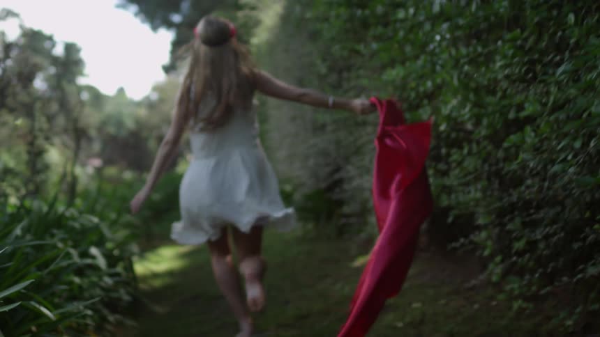 A woman wearing a white dress and holding a length of red material, skipping down grass lined with bushes and plants