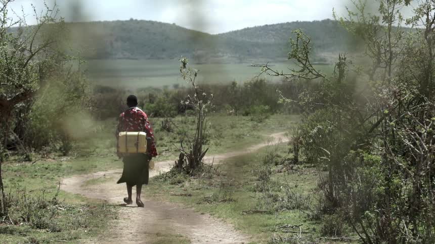 A person with a yellow container on their back walks a dusty path