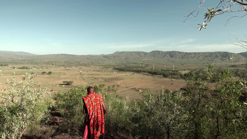 A man wrapped in a red plaid cloth using a walking stick as he descends a trail in a mountainous desert region