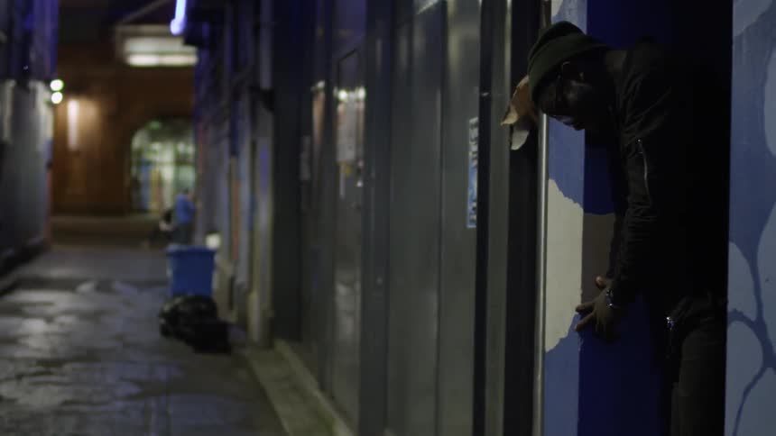 A man in a knit cap stands in a doorway in a narrow alley at night
