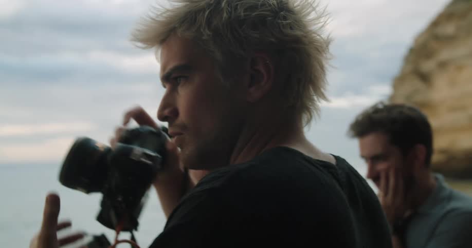 A photographer squints as he focuses and takes a picture on a ridge overlooking the ocean with two men standing beside him
