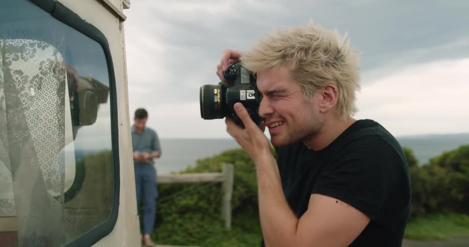 A man standing at the back of a van squints as he aims and focuses his camera