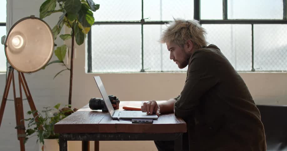 A man sits at a table scrolling on a laptop with a camera beside him and a large light in front of him