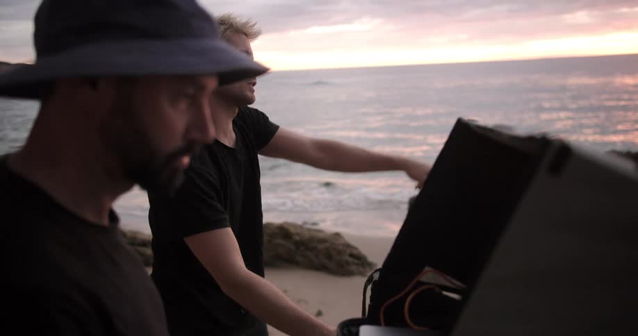 Two men standing on a beach are checking their equipment as one of them issues instructions