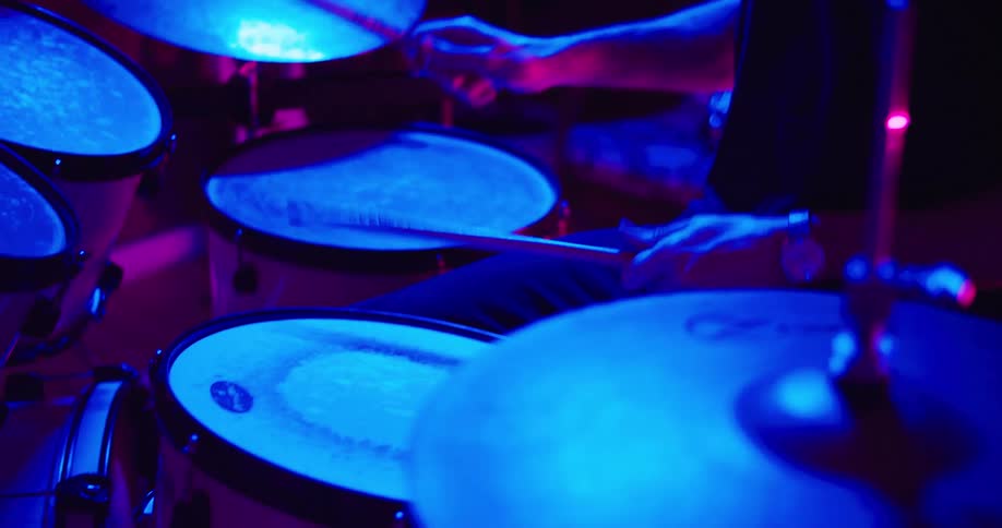 A man drums in a dark room lit with blue and pink lights