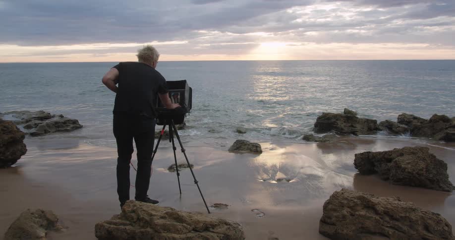 A person stands behind a large commercial video camera pointing at the ocean