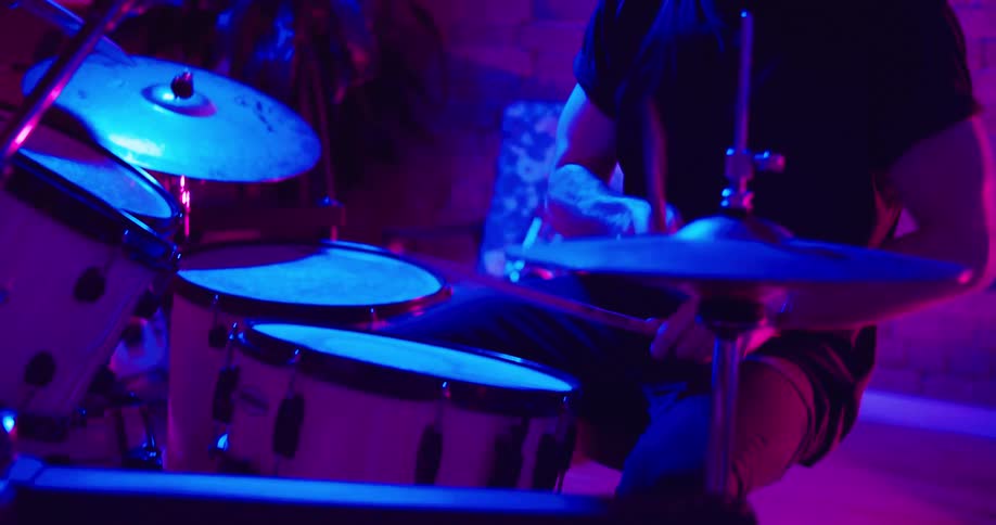 A man plays the drums in a darkened room with pink and blue lights