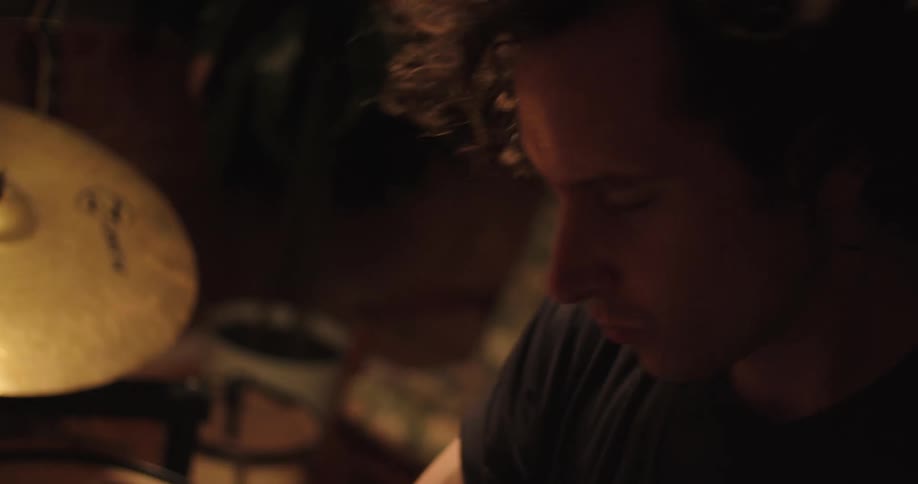 A young man with curly hair energetically plays a set of drums