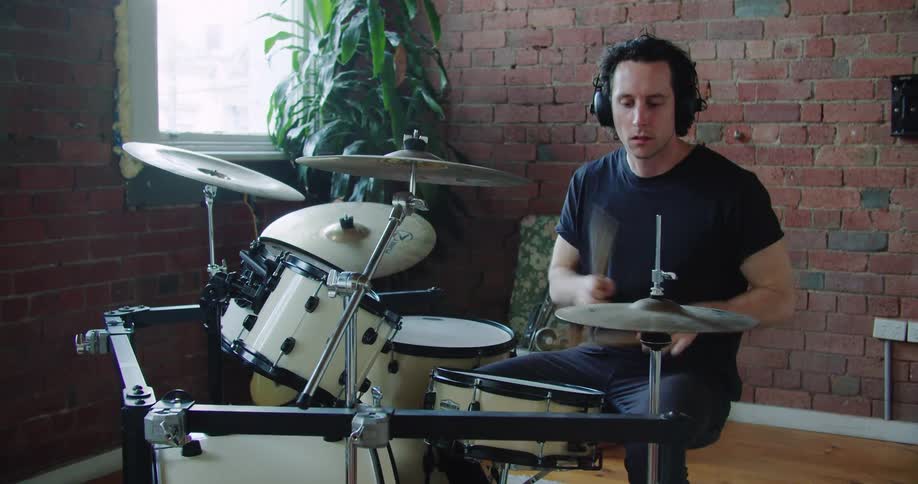A white male wearing headphones is playing the drums in the corner of a room with exposed brick walls