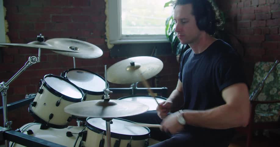 A man wearing headphones plays the drums in a brick walled room with windows