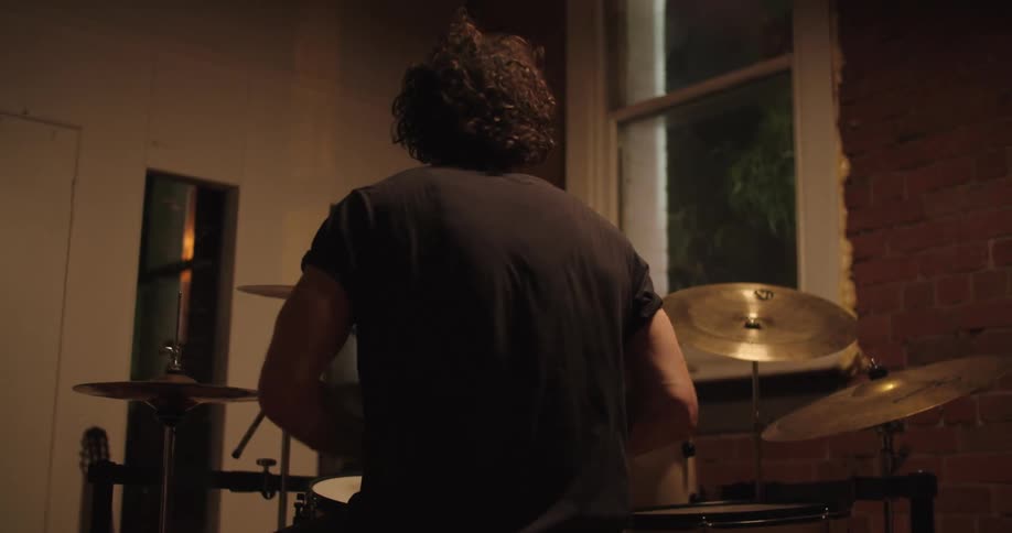 A male with curly hair sits behind a small drum set and plays it with vigor
