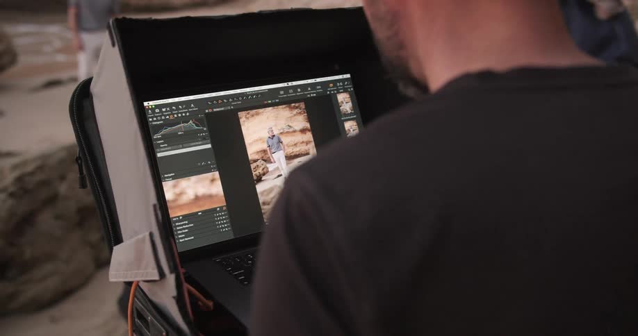 A man uses a laptop to edit a photograph live while the model stands near the ocean
