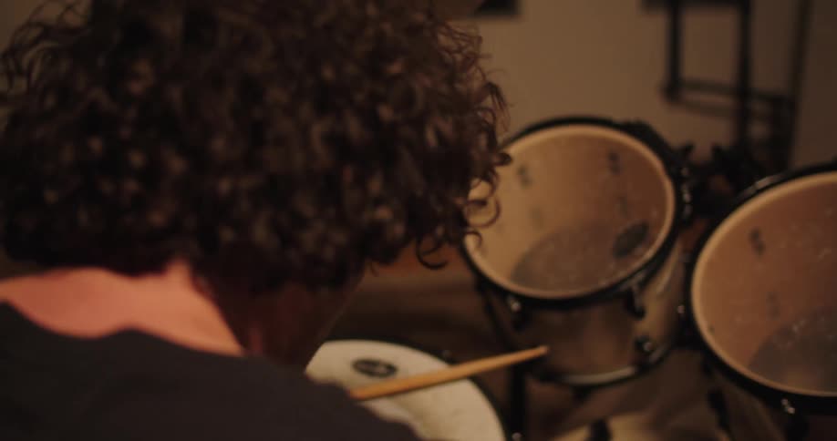 A white man sits at a drum kit motionless before suddenly hitting a drum