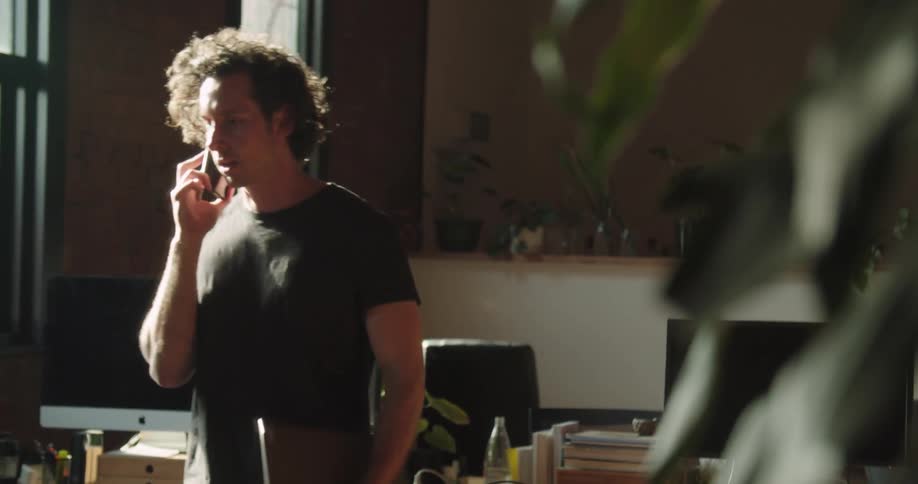 A man paces in his living room while talking on the phone and holding a laptop