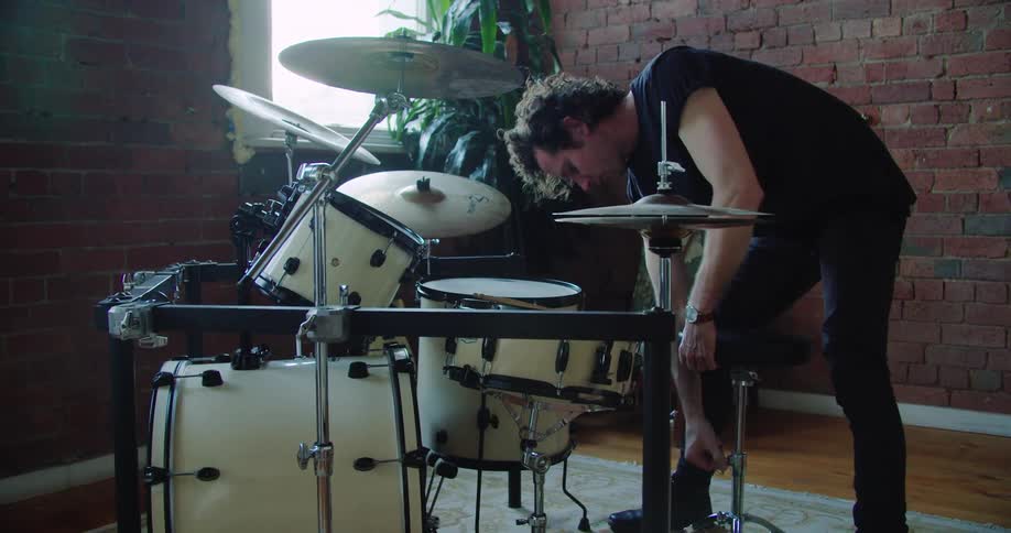 A man adjusts the stool behind his drum set and sits down