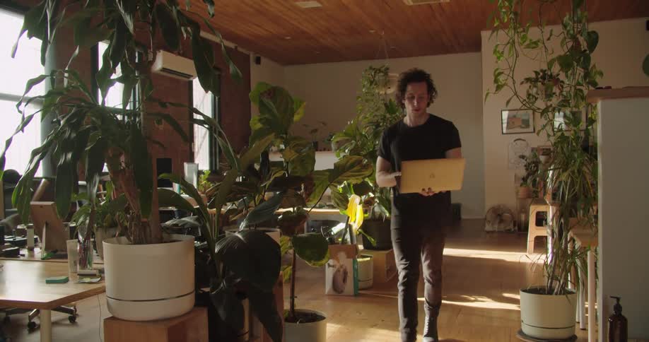 A man walks through a room with lots of plants carrying a laptop