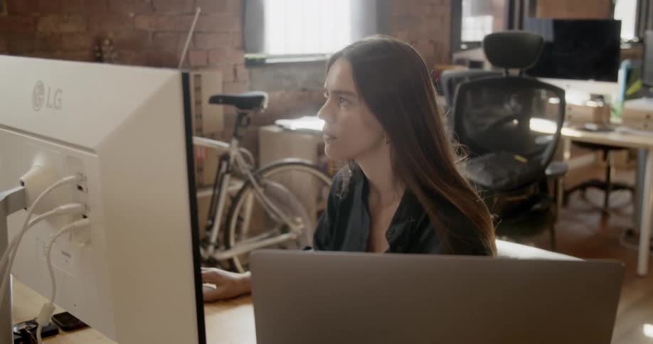 A young woman sits at a computer screen within a cluttered office space