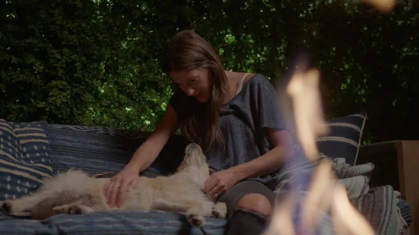 A woman sits outside with her dog and rubs its belly as a fire burns in front of them