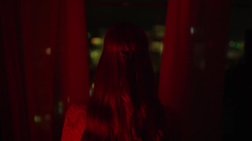 A woman is standing at a large window looking out at the city's lights at night